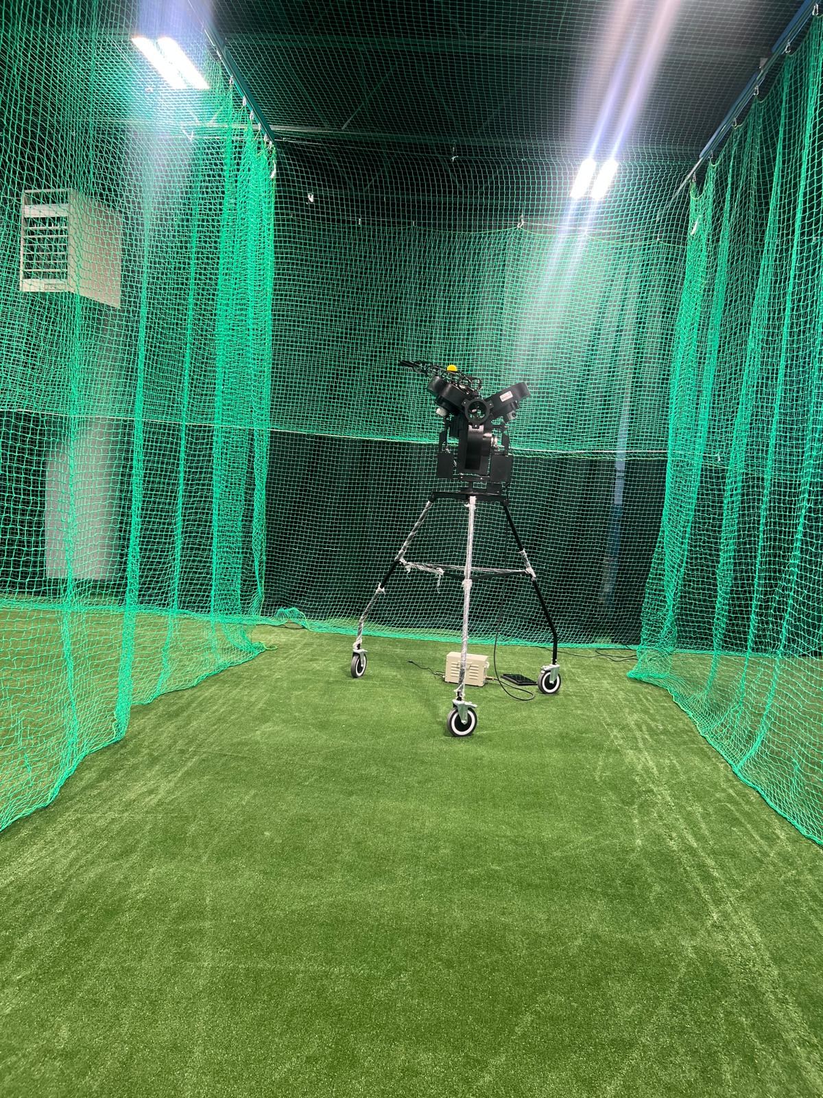 Bowling machine on a wheeled stand at the end of a green-netted practice cage